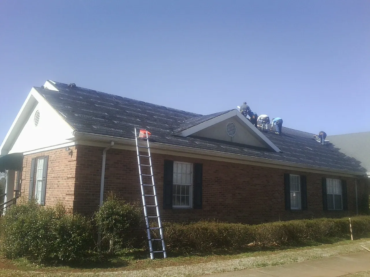Crew tearing shingles off a brick ranch roof with a ladder set — Metal Roof Repair work by our crew in Matthews